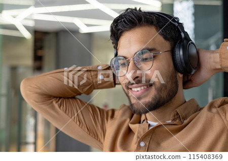 Close-up photo of a smiling young Indian man sitting in the office and resting, hands behind his head, listening to music in headphones and looking ahead. Close-up photo of a smiling young Indian man sitting in the office and resting, hands behind his head, listening to music in headphones and looking ahead. 115408369