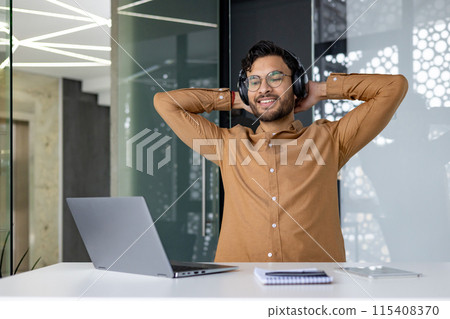 Man in office enjoying music with headphones and laptop in modern workspace. Relaxed employee taking a break from work. Man in office enjoying music with headphones and laptop in modern workspace. Relaxed employee taking a break from work. 115408370