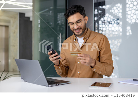 Smiling young male Muslim businessman sitting in the office at the table, holding the phone in his hands and using a credit card, online account. Smiling young male Muslim businessman sitting in the office at the table, holding the phone in his hands and using a credit card, online account. 115408419