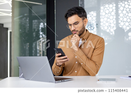 A focused businessman checking his phone while working on a laptop in a modern office setting, portraying concentration and productivity. 115408441