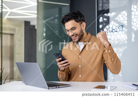 Excited man in a modern office celebrating success by looking at his smartphone and raising his fist in joy. Excited man in a modern office celebrating success by looking at his smartphone and raising his fist in joy. 115408451