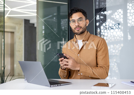 Portrait of a young Indian man in a brown shirt and glasses sitting at a desk in the office, holding a phone and looking at the camera. 115408453