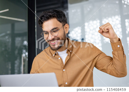 A cheerful man with glasses and a beard celebrates a successful achievement while working on his laptop in a modern, well-lit office. A cheerful man with glasses and a beard celebrates a successful achievement while working on his laptop in a modern, well-lit office. 115408459