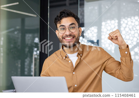 Close-up photo of a young Muslim man who is in the office and shows the camera a victory gesture with his hand, impressed with success and achievement, looking at the camera with a smile. Close-up photo of a young Muslim man who is in the office and shows the camera a victory gesture with his hand, impressed with success and achievement, looking at the camera with a smile. 115408461