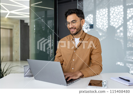 Indian young man studying and working in a modern office, sitting at a desk, smiling and typing on a keyboard with concentration. Indian young man studying and working in a modern office, sitting at a desk, smiling and typing on a keyboard with concentration. 115408480