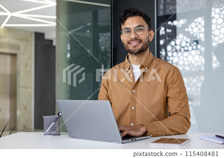 Portrait of a young smiling Indian male student and office worker sitting at a desk, working and studying on a laptop. Looks confidently into the camera. Portrait of a young smiling Indian male student and office worker sitting at a desk, working and studying on a laptop. Looks confidently into the camera. 115408481