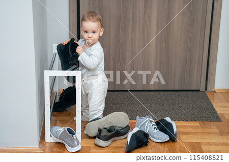 Child in hallway playing with adult boots. Concept of toddler mischief and playful discovery 115408821