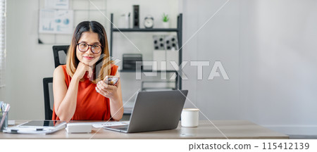 A woman in an orange shirt is sitting at a desk with a laptop and a cell phone. She is looking at her cell phone and she is focused on something A woman in an orange shirt is sitting at a desk with a laptop and a cell phone. She is looking at her cell phone and she is focused on something 115412139