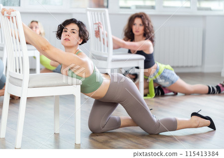 Positive young woman posing on knees near chair on floor in dance studio during group class of strip plastic and dance in high heels in dance studio 115413384