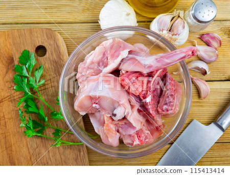 Glass bowl with pieces of raw rabbit on table with greens and condiments 115413414