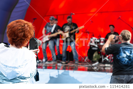 Woman capturing memories with a video camera at a vibrant concert with a striking red backdrop Woman capturing memories with a video camera at a vibrant concert with a striking red backdrop 115414281