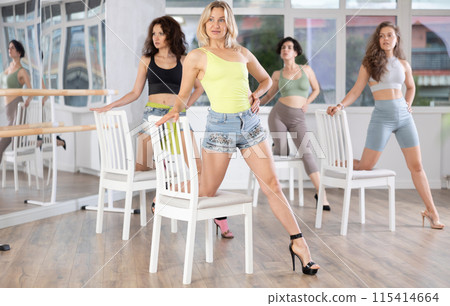 Active female dancer with group of women of different ages practicing strip plastic movements near chair in training hall during dancing classes 115414664