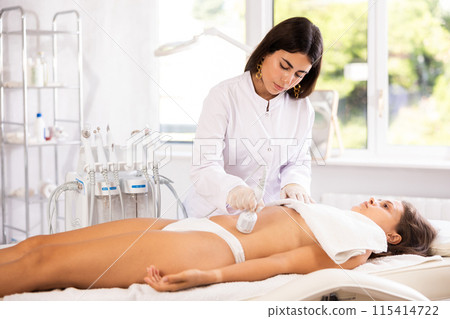 Cosmetologist performing cryotherapy procedure with cooling hammer on female client belly 115414722