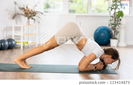 Young woman yoga trainer performing downward dog pose on black rug 115414873