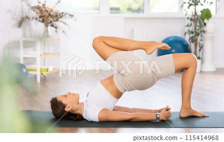 Young woman yoga trainer performing one legged bridge pose on black rug 115414968