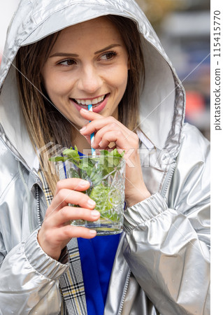 Young woman happily sips drink with mint, wearing stylish silver jacket, creating modern look Young woman happily sips drink with mint, wearing stylish silver jacket, creating modern look 115415770