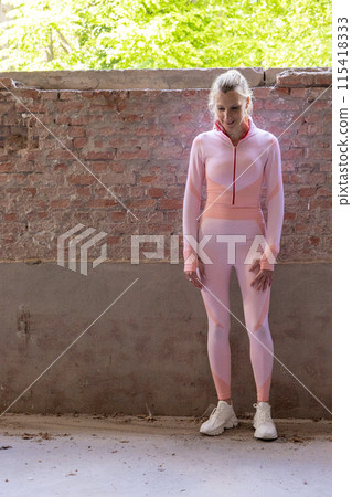 A woman wearing pink sportswear is seen standing next to a brick wall in an urban environment 115418333