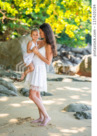 Young woman mother with a little daughter in white dresses on seashore in the shade of trees and palms Young woman mother with a little daughter in white dresses on seashore in the shade of trees and palms 115420194