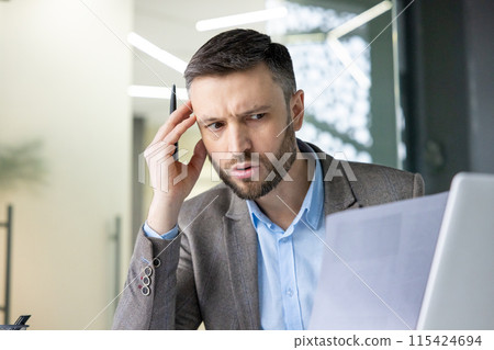 Businessman in office looking confused while reading a document and working on a laptop, holding a pen in hand. 115424694