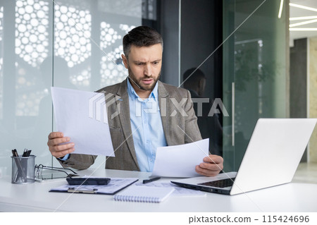 Businessman examining papers, multi-tasking at an office desk with a laptop, looking concerned or focused. Businessman examining papers, multi-tasking at an office desk with a laptop, looking concerned or focused. 115424696