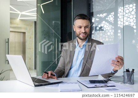 Smiling businessman holding a document while working in a modern office. Professional environment with a laptop, papers, and a pen. Smiling businessman holding a document while working in a modern office. Professional environment with a laptop, papers, and a pen. 115424697