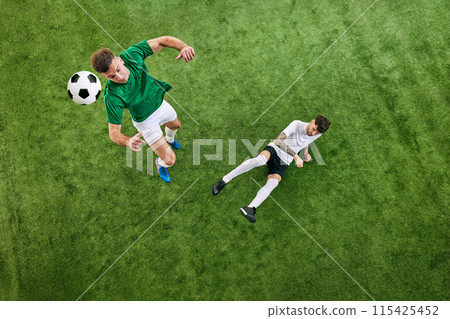 High-angle view. Two soccer players in action on green lush playground. Manin green aims to kick ball, while opponent stays alert and ready to defend. 115425452