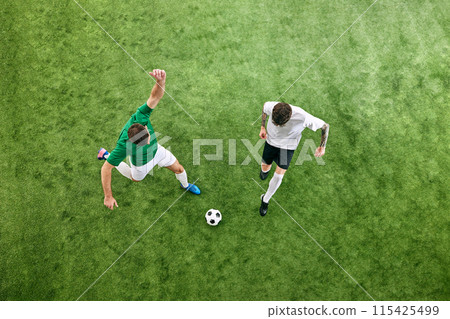 Aerial view. Two players in white and green uniforms battle for the ball on green lush playground. Aerial view. Two players in white and green uniforms battle for the ball on green lush playground. 115425499
