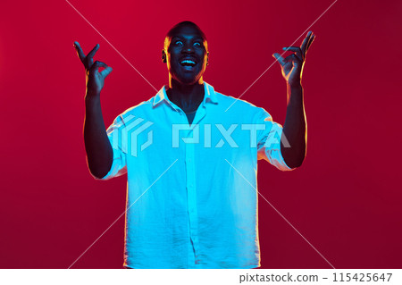 Portrait of emotional African-American man in white shirt raising hands upwards with excited face against red background in neon light Portrait of emotional African-American man in white shirt raising hands upwards with excited face against red background in neon light 115425647