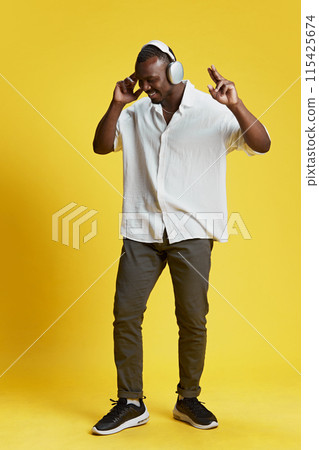 Full-length portrait of African-American man in casual clothes listening to music in headphones and dancing against bright yellow background 115425674