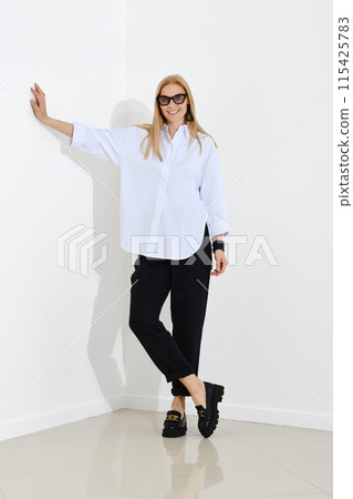 Woman in white shirt and trousers leans her hand against the wall while standing in a bright room 115425783