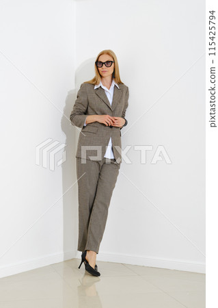 Stylish professional woman in checkered suit leaning against white wall 115425794