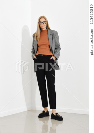 Stylish woman wearing checkered blazer, turtle neck and trousers standing against white office wall Stylish woman wearing checkered blazer, turtle neck and trousers standing against white office wall 115425819