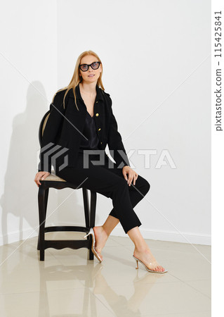 Cute woman in a black jacket and tight trousers sits in profile on a chair against a white wall 115425841