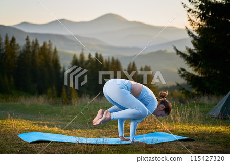 Woman practicing yoga outdoors in the mountains in a serene, natural setting. Female performing yoga pose on blue mat, with backdrop of beautiful mountain landscape at sunrise or sunset. Woman practicing yoga outdoors in the mountains in a serene, natural setting. Female performing yoga pose on blue mat, with backdrop of beautiful mountain landscape at sunrise or sunset. 115427320