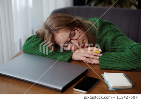 A woman in a green sweater is sleeping by a laptop on a desk 115427597