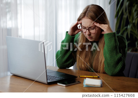 A woman in a green sweater is seated at a desk with a laptop 115427608