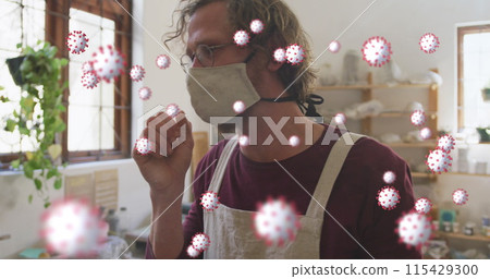 Image of coronavirus cells floating over caucasian man in face mask coughing at pottery workshop 115429300