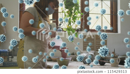 Image of covid 19 cells over woman working in pottery studio wearing face mask 115429313