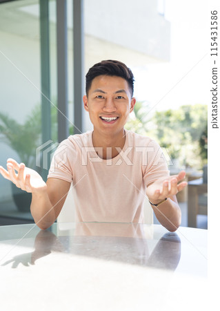 Young Asian man gestures while sitting at a table on a video call Young Asian man gestures while sitting at a table on a video call 115431586