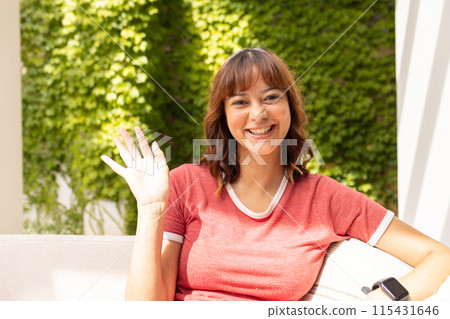 Young biracial woman waves cheerfully on video call, seated outdoors 115431646