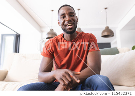 African American man in an orange shirt smiles warmly, seated on a couch on video call African American man in an orange shirt smiles warmly, seated on a couch on video call 115432371