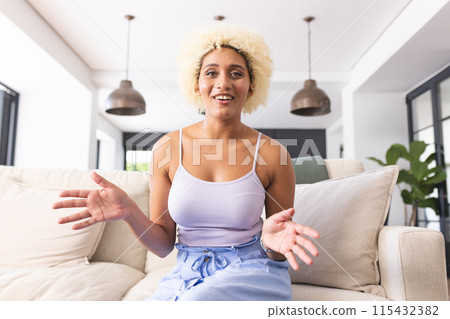 Young biracial woman with curly blonde hair sits on couch, gesturing while talking on a video call 115432382