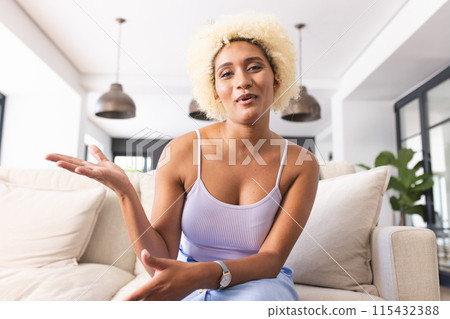 Young biracial woman with curly blonde hair gestures while talking on a video call, seated on a sofa 115432388