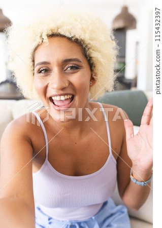 Young biracial woman with curly blonde hair smiles joyfully, waving at the camera on a video call Young biracial woman with curly blonde hair smiles joyfully, waving at the camera on a video call 115432397