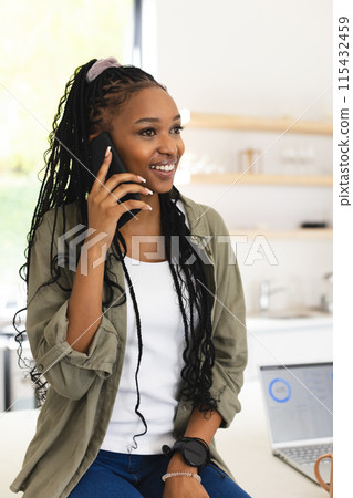 Young African American woman chats on her phone, a laptop open before her 115432459