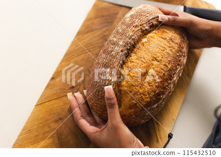 Hands slice a large loaf of bread on a wooden cutting board with copy space 115432510