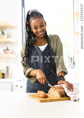 A young African American woman slices bread in a bright kitchen 115432517