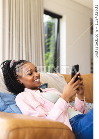 A young African American woman lounges on a sofa, smiling at her phone, with copy space 115432633