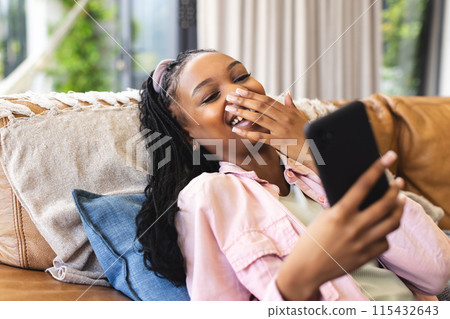 Young African American woman laughs while looking at her phone, reclining on a couch 115432643