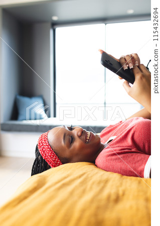 A young African American woman lies on a bed, smiling while holding a smartphone, with copy space A young African American woman lies on a bed, smiling while holding a smartphone, with copy space 115432694